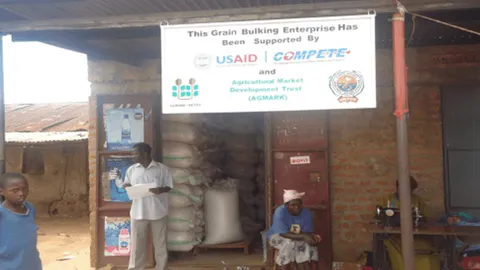Grain bulking enterprise storefront with a banner crediting USAID, COMPETE, and AGMARK support