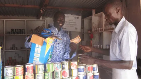 Two men completing a transaction at a market stall stocked with canned goods