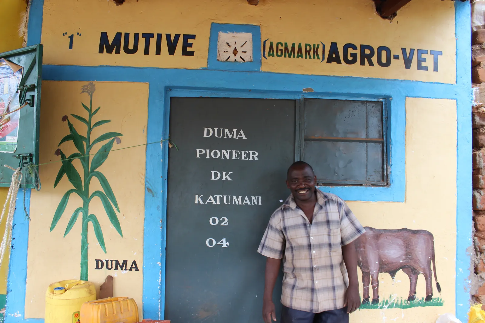 Smiling agro-dealer standing in front of his hand-painted Mutive Agro-Vet shopfront featuring AGMARK branding and seed-variety signage