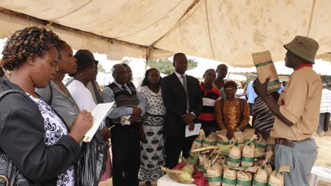 Vendor showing a seed packet to a group gathered under a tent, with sacks of produce on the table