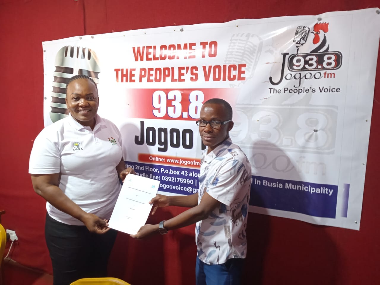 Man and Woman exchange document indoors with vibrant banner backdrop for AGMARK and Jogoo FM Busia cross-border trade partnership on Uganda trade radio partnership